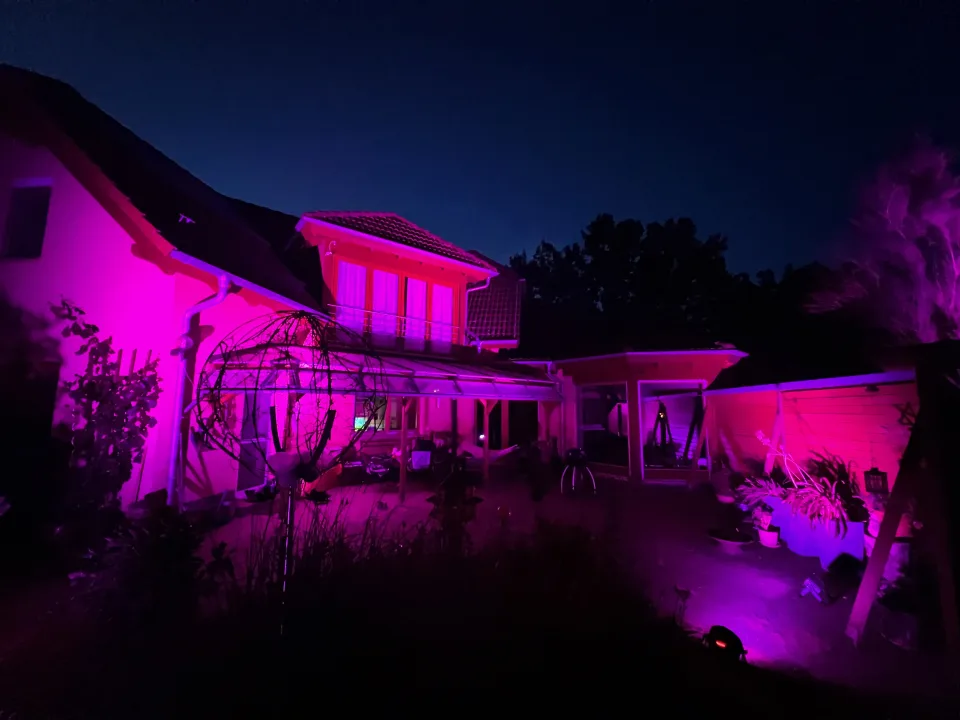 In Magenta angestrahlte Hausfassade mit Ambientebeleuchtung im Garten zur blauen Stunde